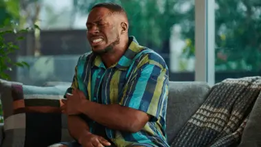 A man in a plaid shirt sits on a sofa, grimacing in pain while clutching his left shoulder. The room is softly lit with greenery outside the windows.